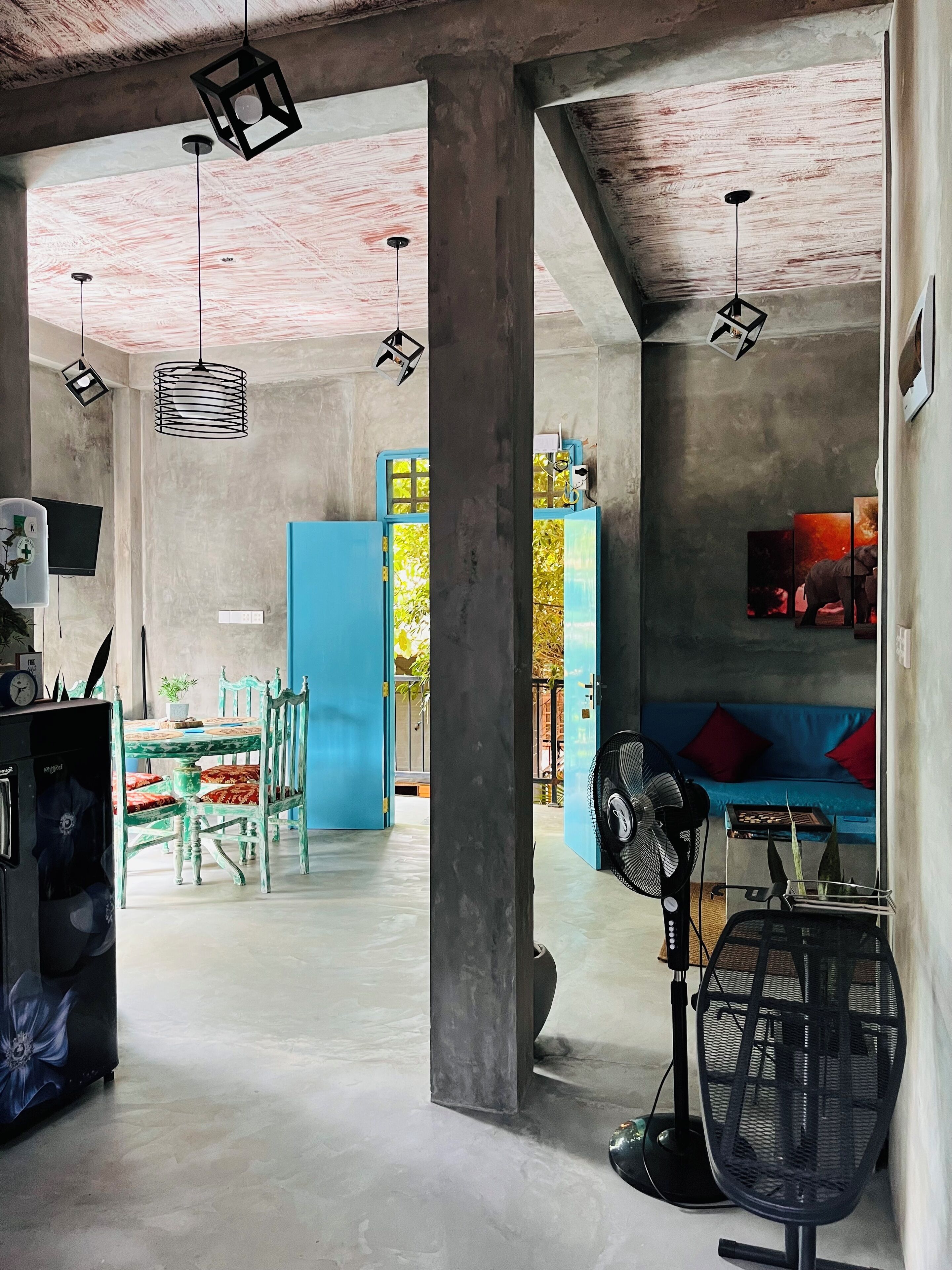 A spacious room with a high ceiling, exposed concrete beams, a blue sofa, a dining table with chairs, and a large fan.