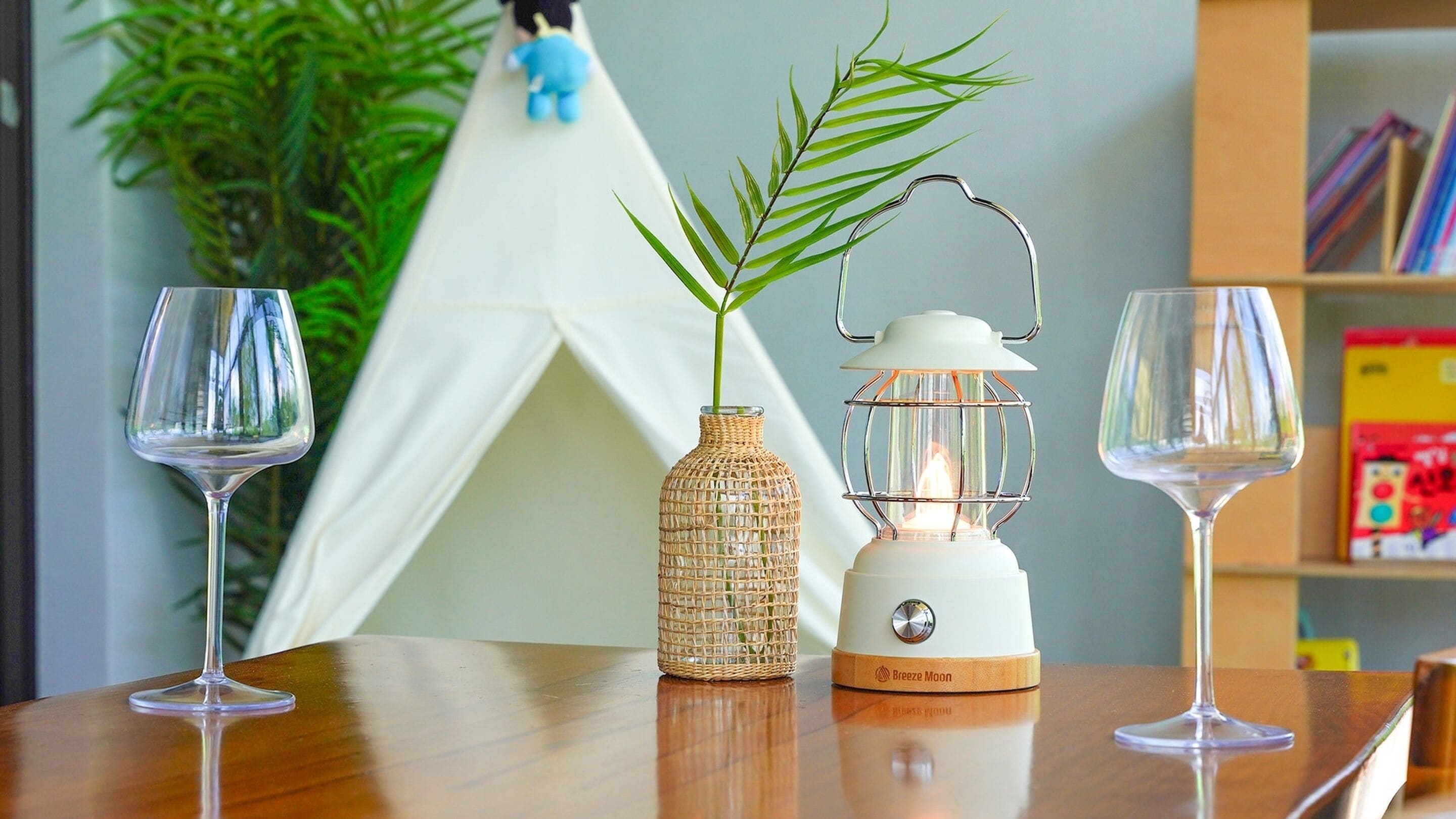 A wooden table with two wine glasses, a woven basket with a plant, and a white lantern on a shelf.