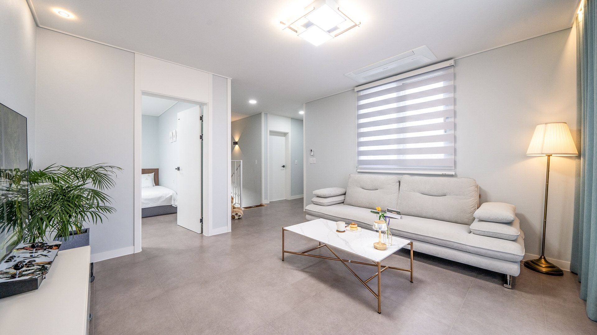 Ein modernes Wohnzimmer mit einer Couch, einem Couchtisch und einer Lampe. Im angrenzenden Raum befindet sich ein Bett und in der Ecke steht eine Pflanze.