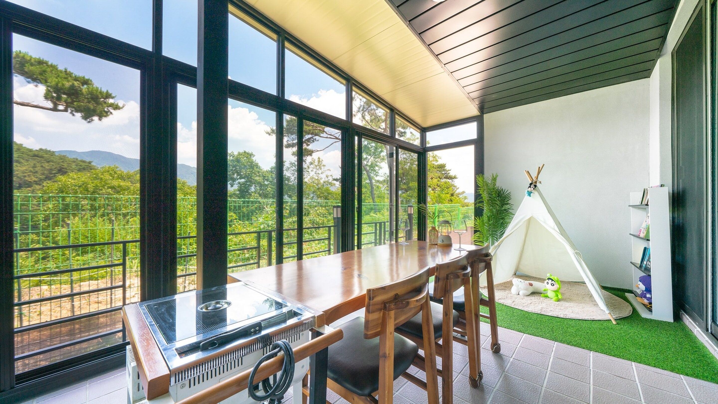 A dining area with a large table, chairs, and a grill. There is a view of greenery and mountains through large windows. A teepee and a bookshelf are visible in the background.