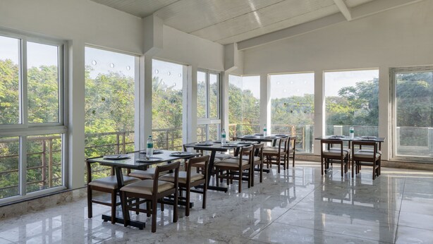 Un comedor espacioso con amplias ventanas que ofrecen vistas a abundante vegetación. Las mesas están ordenadas con sillas, y el suelo está cubierto de baldosas.