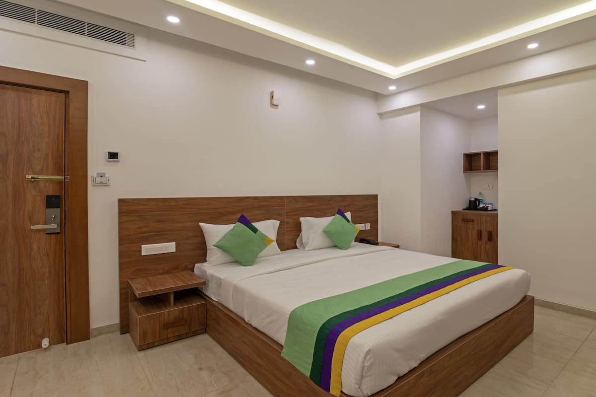 A modern hotel room with a wooden headboard, a large bed with a green and yellow striped blanket, a nightstand, a wooden cabinet, and a small kitchenette area.