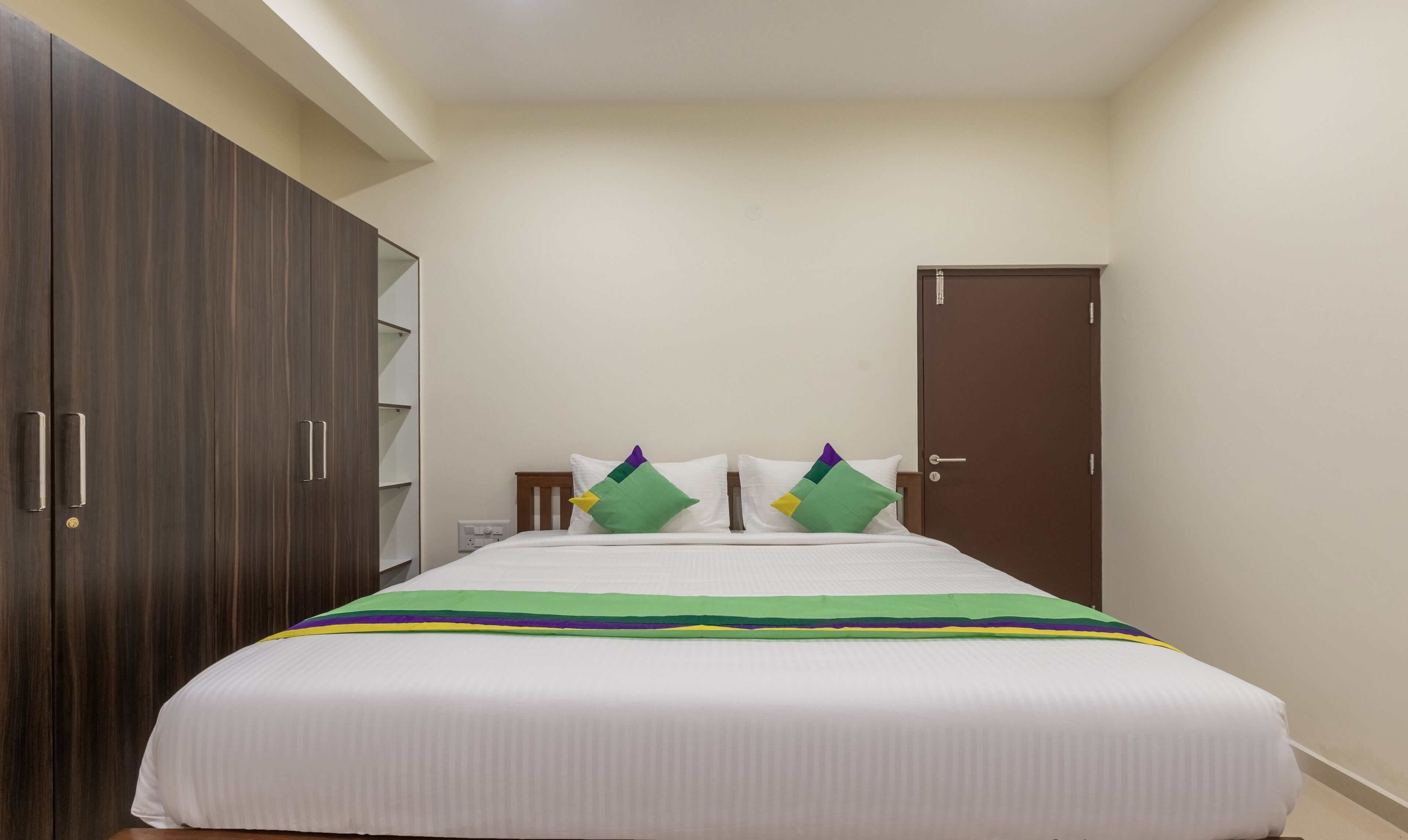 A bedroom with a large wardrobe, a bed with a striped bedspread, and two pillows with green and purple accents.