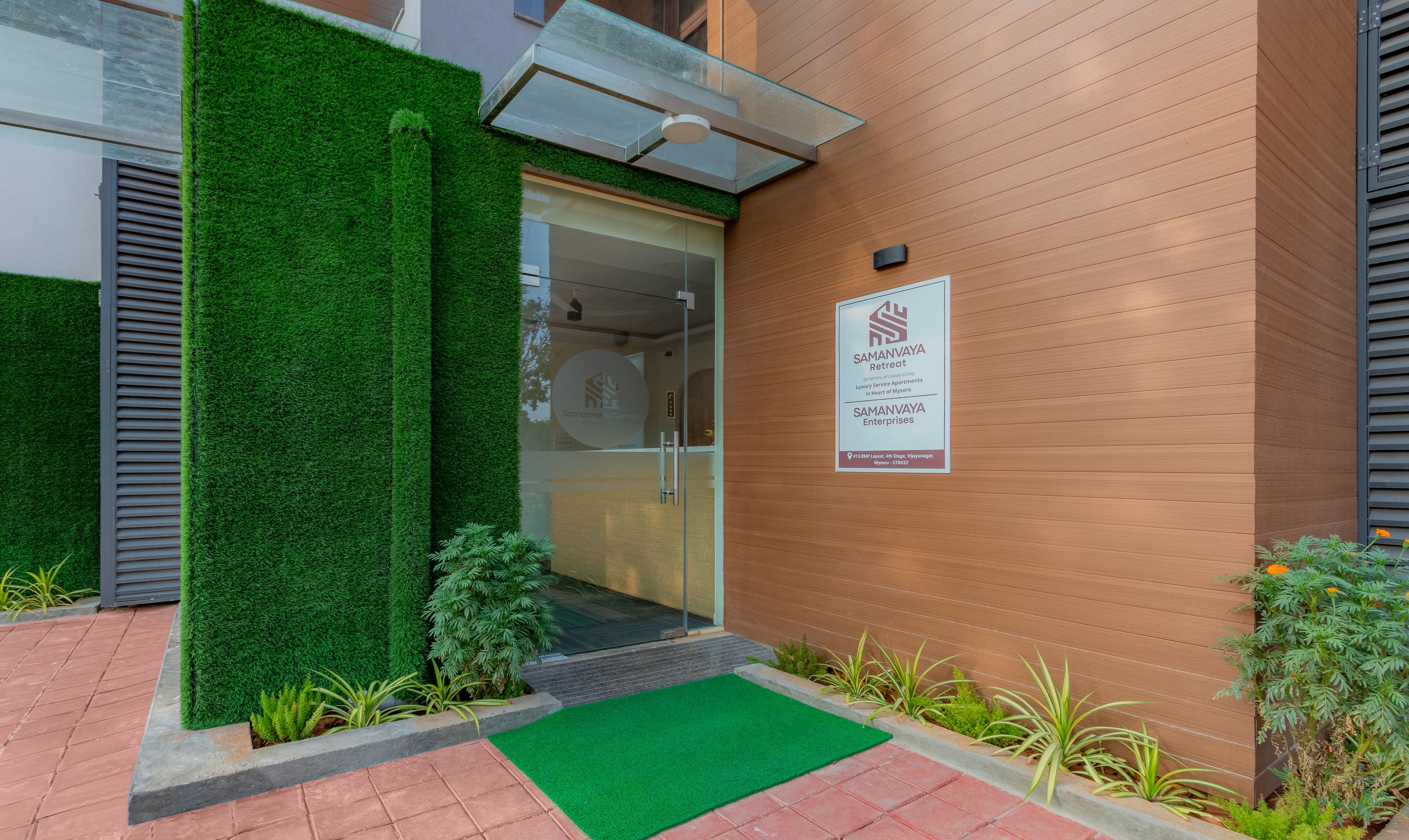 A modern building entrance with a green artificial grass mat, a glass door, and a signboard for Samanvaya Retreat.