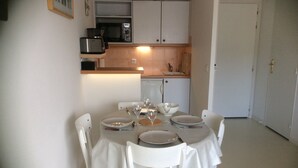 Uma sala de jantar com mesa posta para quatro pessoas, cadeiras brancas e uma cozinha ao fundo.