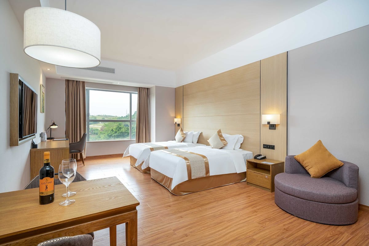 A hotel room with two beds, a desk with a bottle of wine and a glass, a round armchair, and a large window with a view.