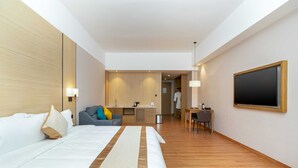 Quarto de hotel com uma cama grande, uma escrivaninha, uma cadeira, uma televisão e um espaço com cozinha compacta.