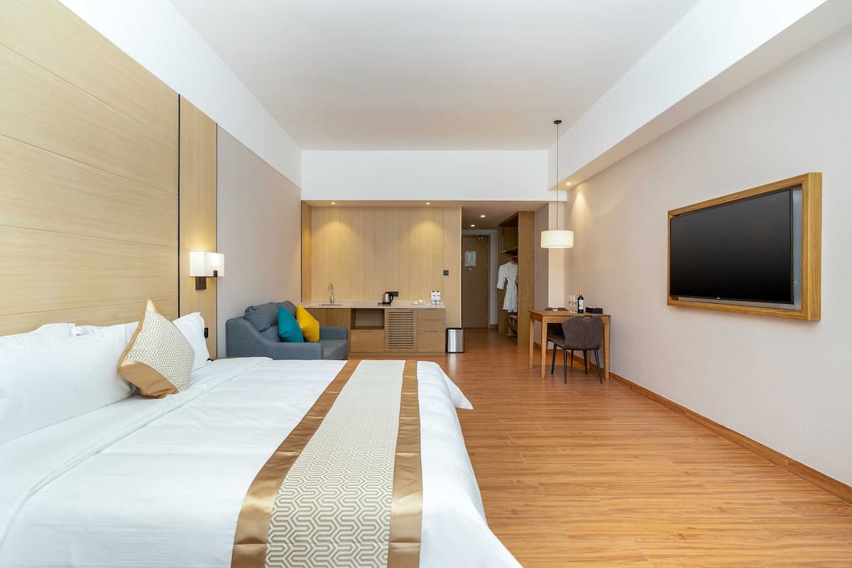 A hotel room with a large bed, a desk, a chair, a TV, and a kitchenette area.
