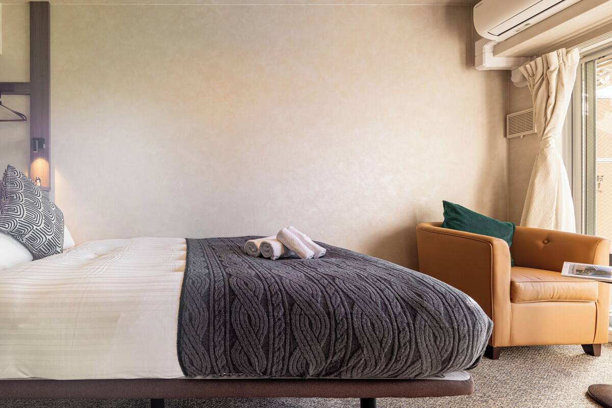 Un lit bien fait, recouvert d’une couverture grise, d’un coussin à motifs et d’une couette blanche. On y trouve une petite table avec un magazine, une chaise et une fenêtre avec des rideaux.