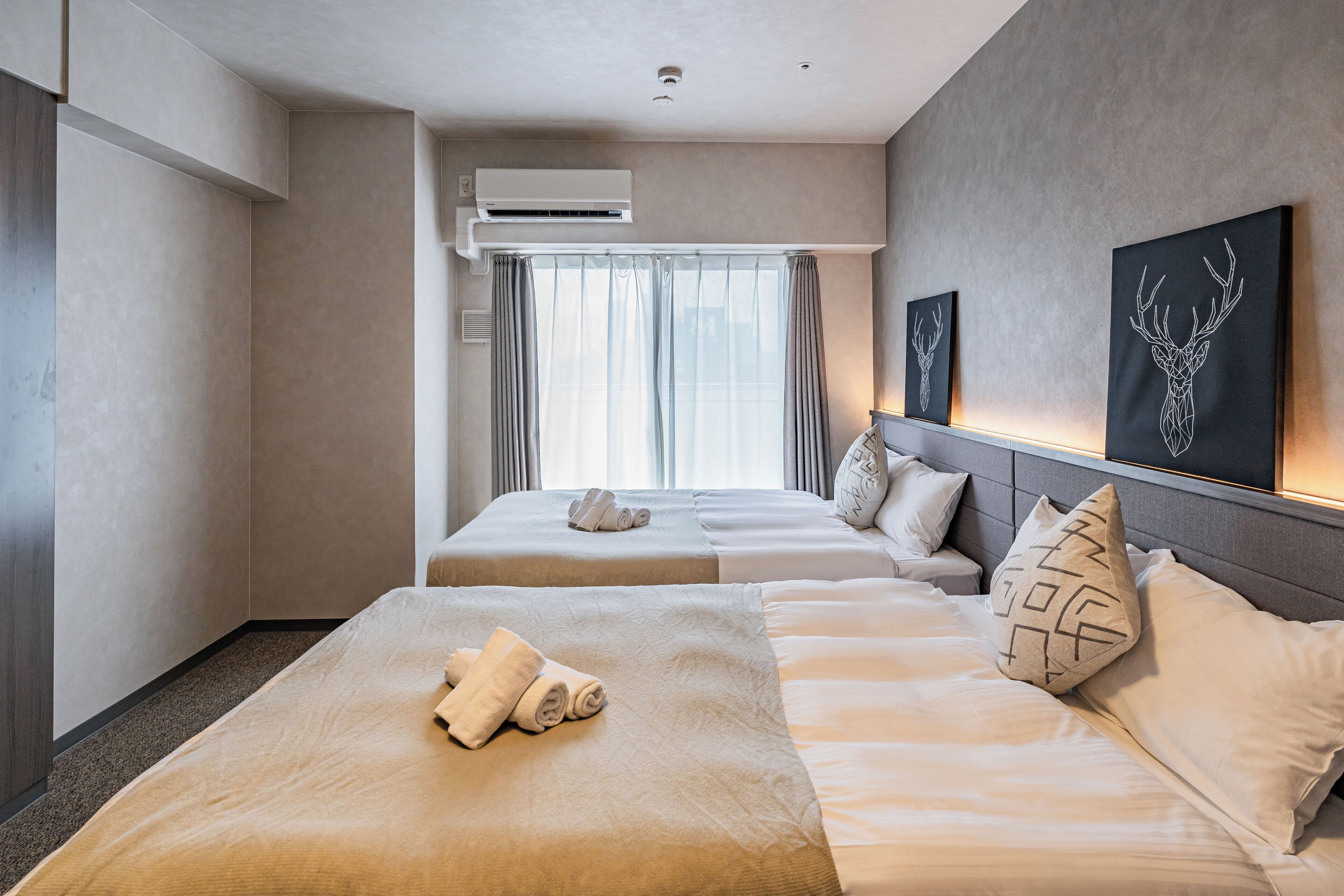 A hotel room with two beds, a large window with curtains, an air conditioning unit, and artwork on the wall.