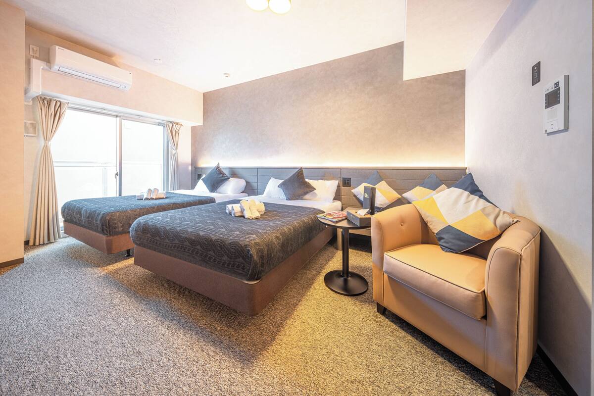 Une chambre d’hôtel avec deux lits, un canapé, une petite table et un luminaire au plafond.
