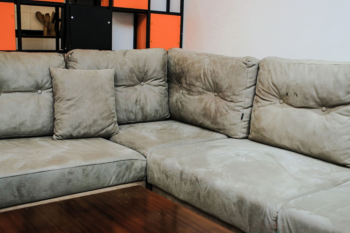 A beige sectional sofa with tufted cushions, positioned in a room with a wooden floor and a black and orange wall panel.