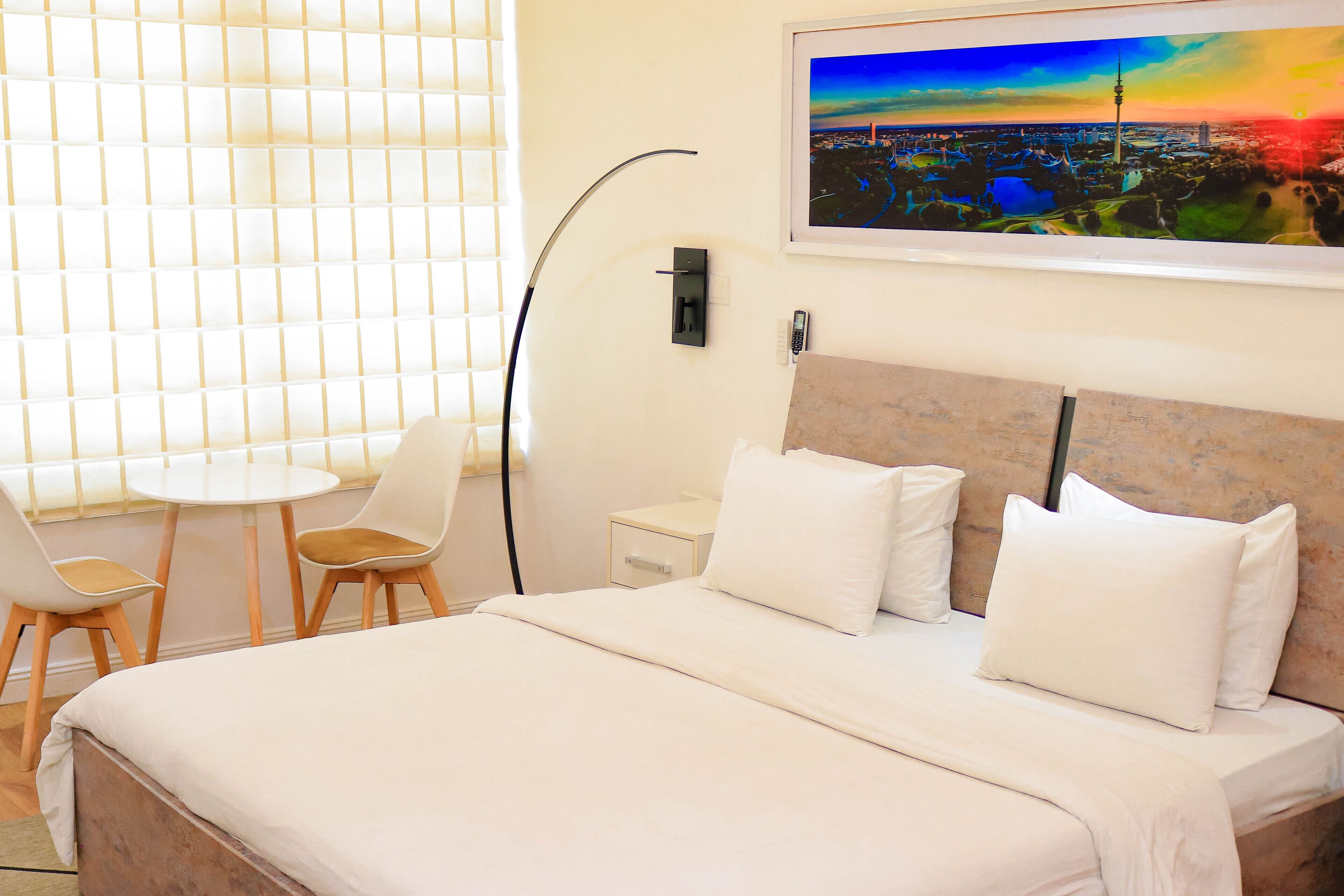 A hotel room with a bed, two chairs, a desk, a lamp, and a painting of a cityscape.