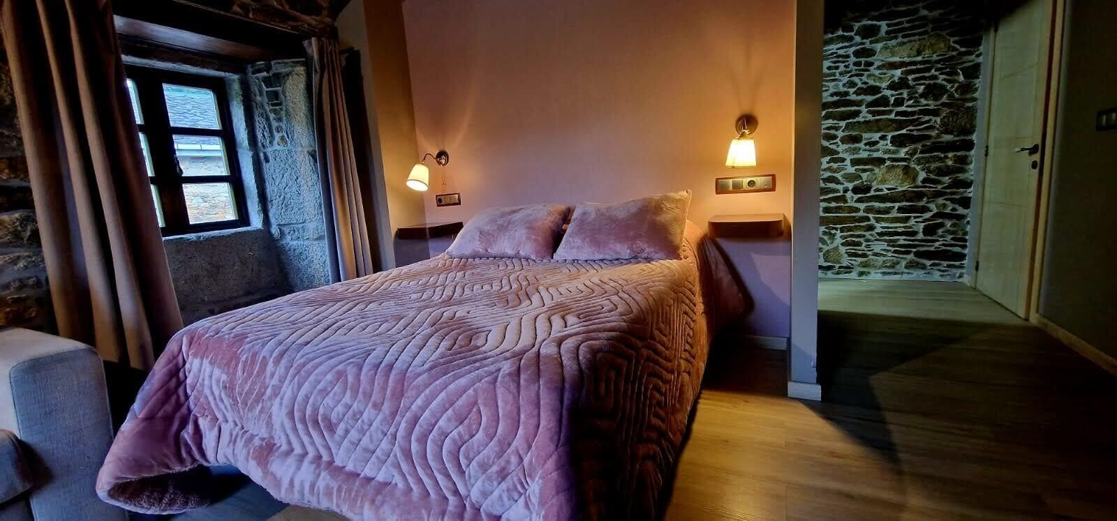 A bedroom with a bed, two bedside lamps, a stone wall, and a window.