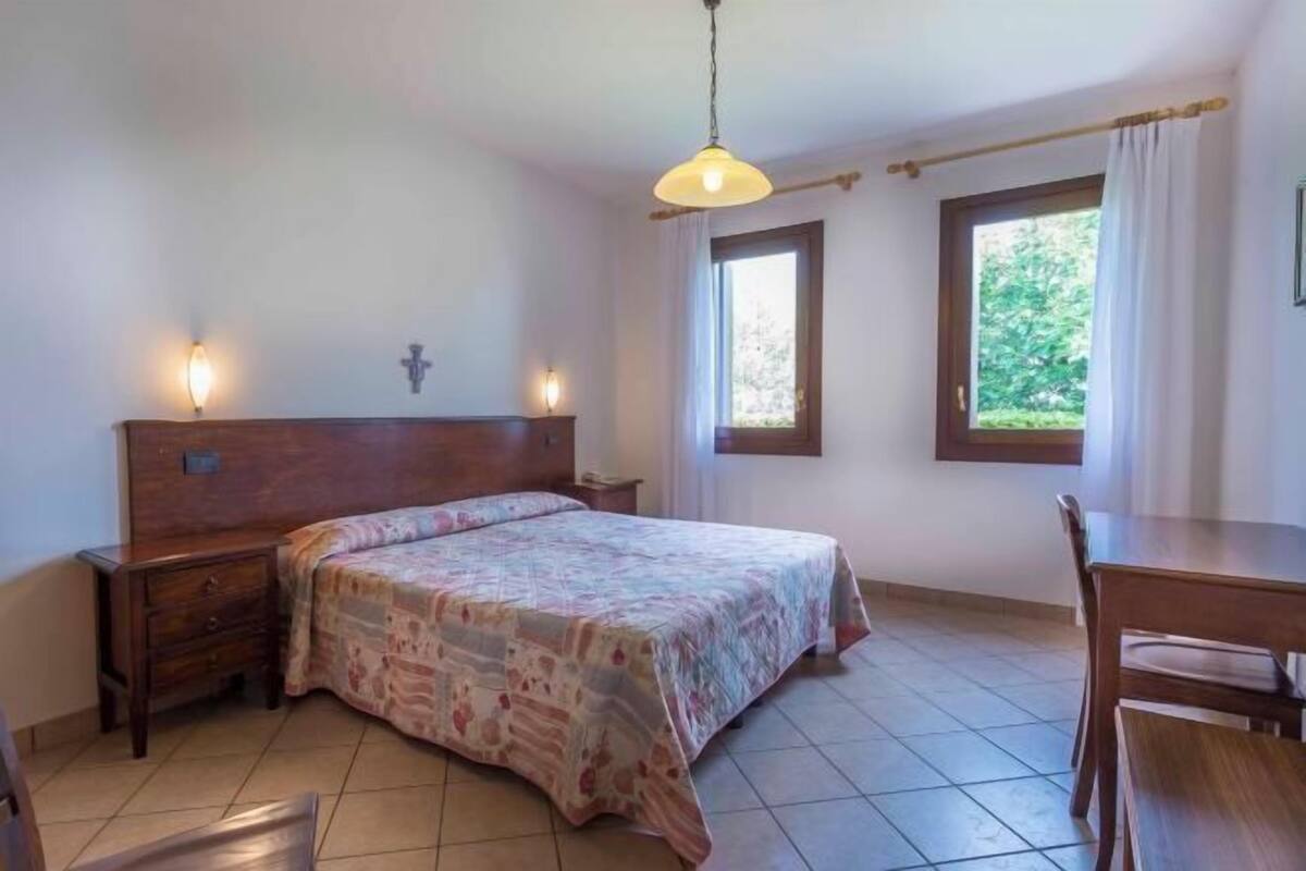 A bedroom with a bed, bedside tables, a wooden headboard, two windows with curtains, and a hanging light fixture.