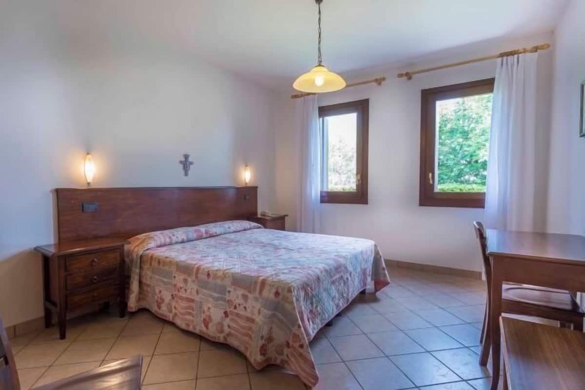 A bedroom with a bed, bedside tables, a wooden headboard, two windows with curtains, and a hanging light fixture.