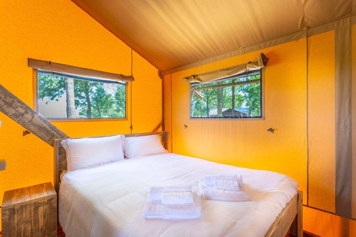 A single bed with white linens and towels, two windows with curtains, and a wooden headboard.