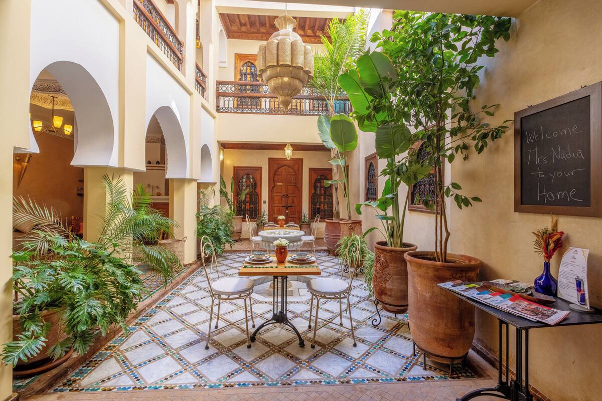 A courtyard with a tiled floor, a central table with chairs, potted plants, and a sign welcoming guests.