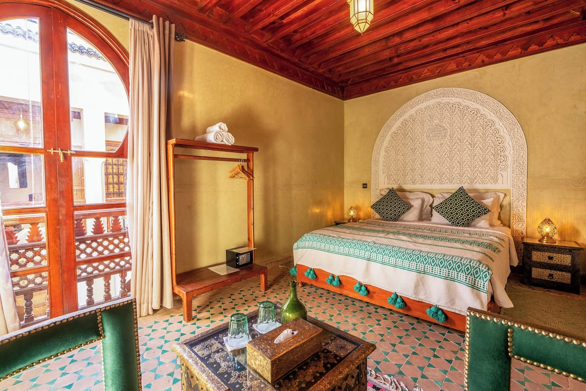 A room with a bed, a wooden chair, a table with a vase, and a patterned floor.
