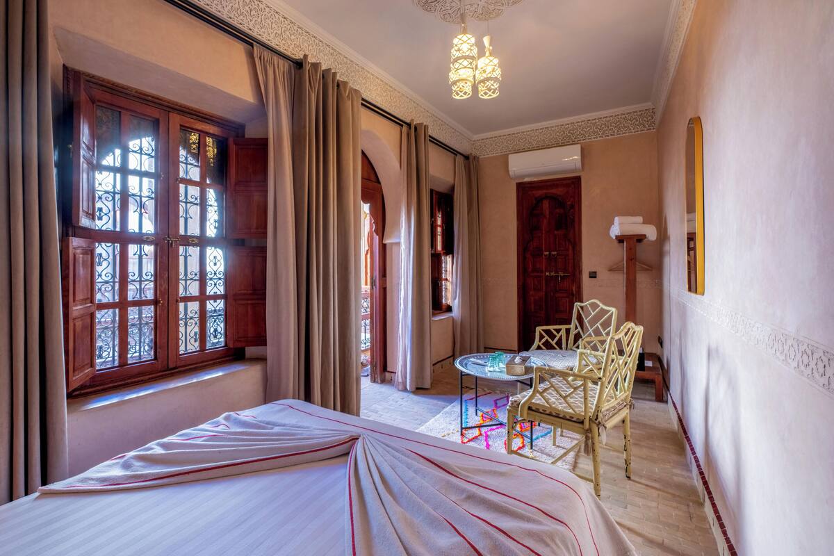 A hotel room with a bed, a small table with chairs, a window with curtains, and a chandelier.