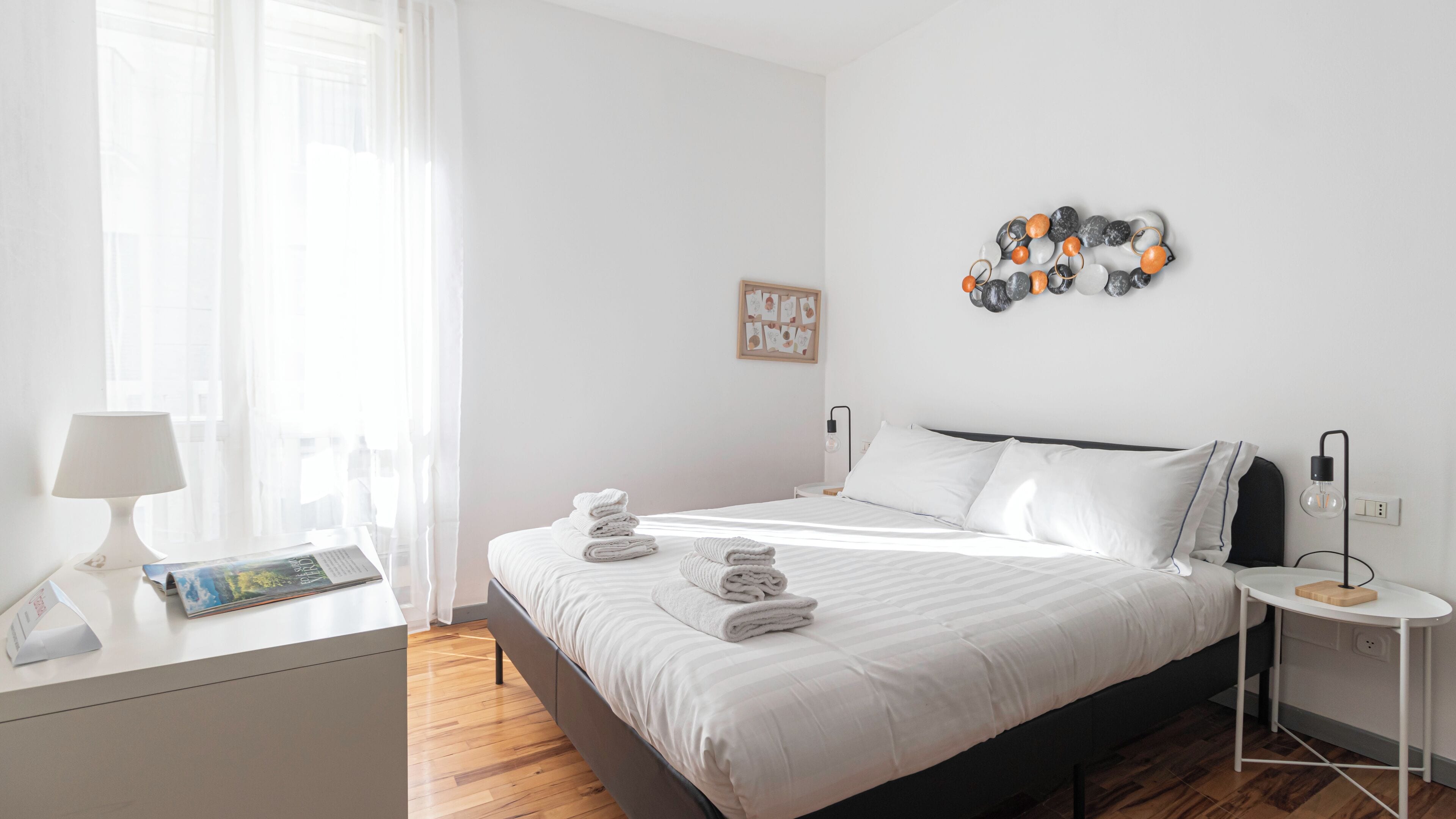 A modern bedroom with a neatly made bed, a bedside table with a lamp, and a wall-mounted light fixture.