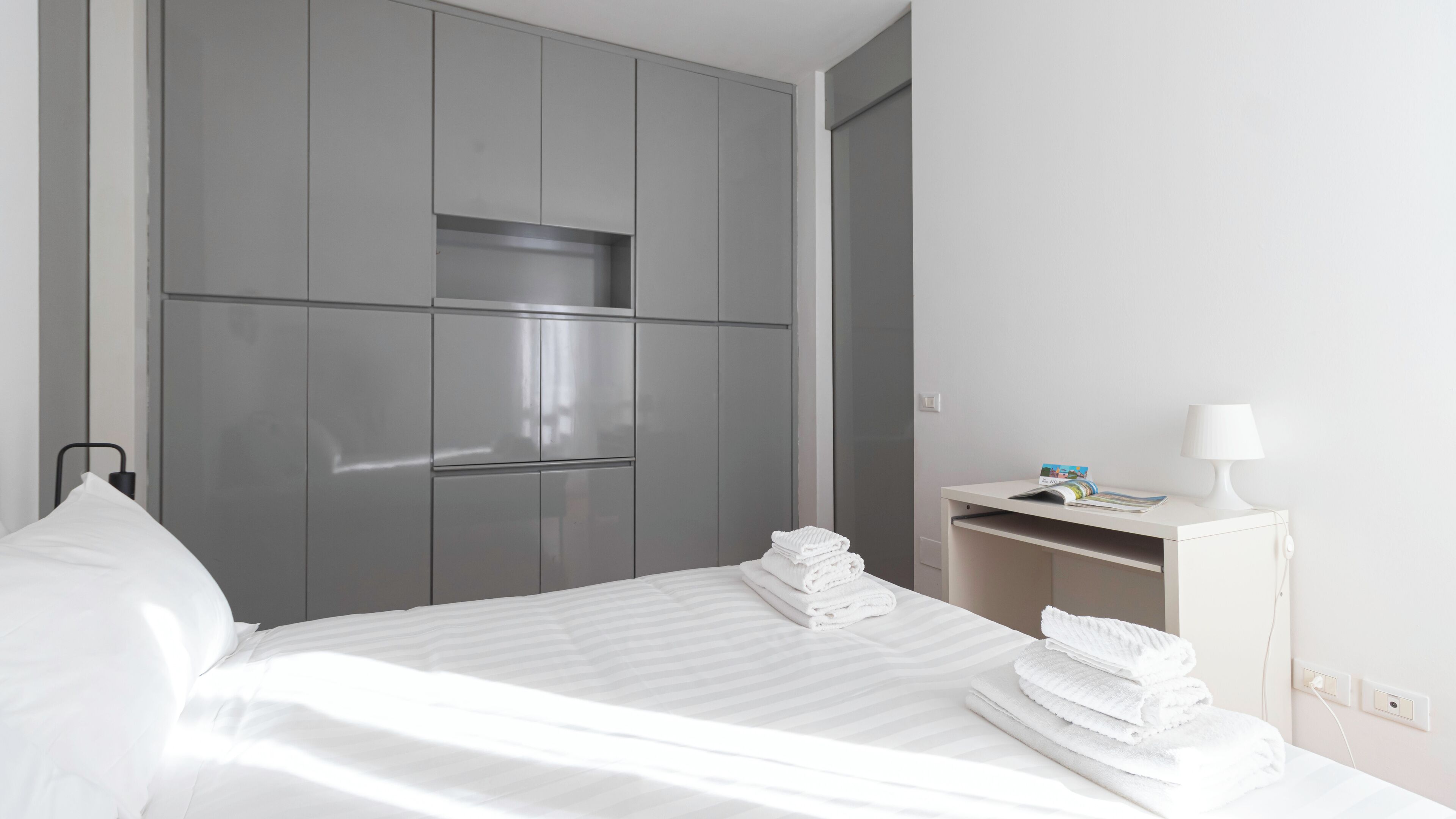 A hotel room with a large bed, grey wardrobe, and a bedside table with a lamp and a magazine.
