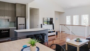A modern kitchen with stainless steel appliances, a dining area with a wooden table and chairs, and a living space with a TV and a staircase.