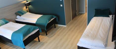 A hotel room with two single beds, each with a teal bedspread and white linens. There is a bedside table with a lamp on one side and a wardrobe on the other. The room has a wooden floor and teal walls.