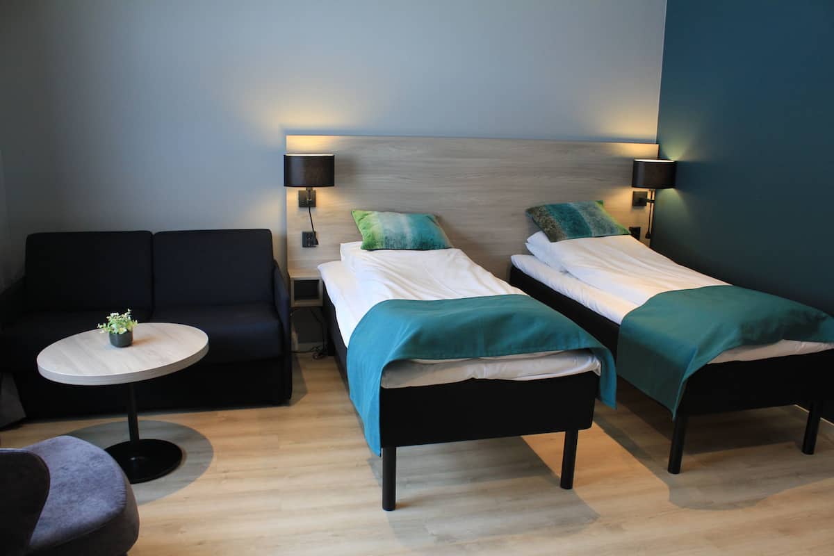 Quarto de hotel com duas camas de solteiro, uma mesa redonda pequena com uma planta, um sofá e duas luminárias de cabeceira.