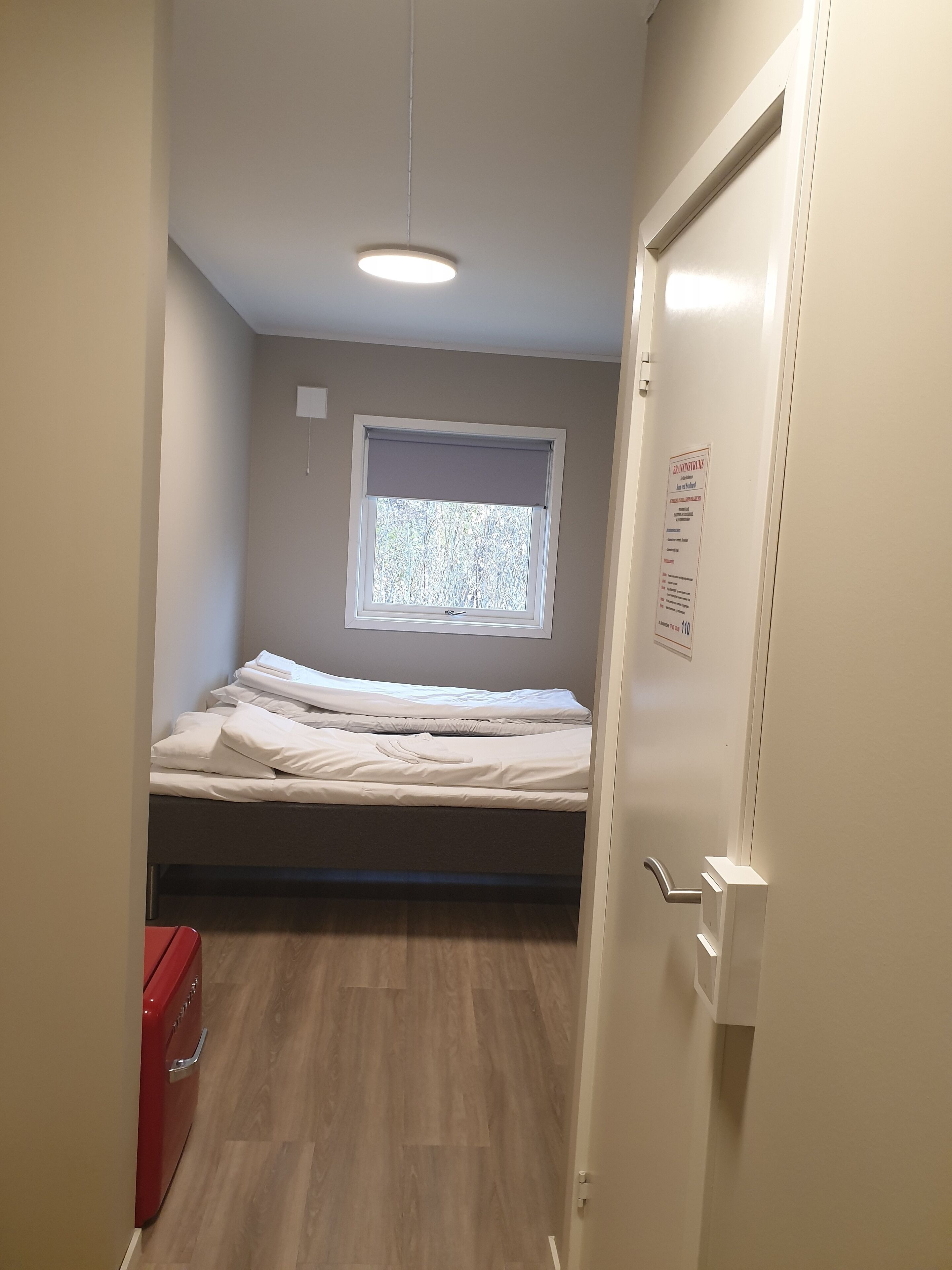 A small, single-bed room with a window, a red suitcase, and a door with a sign.