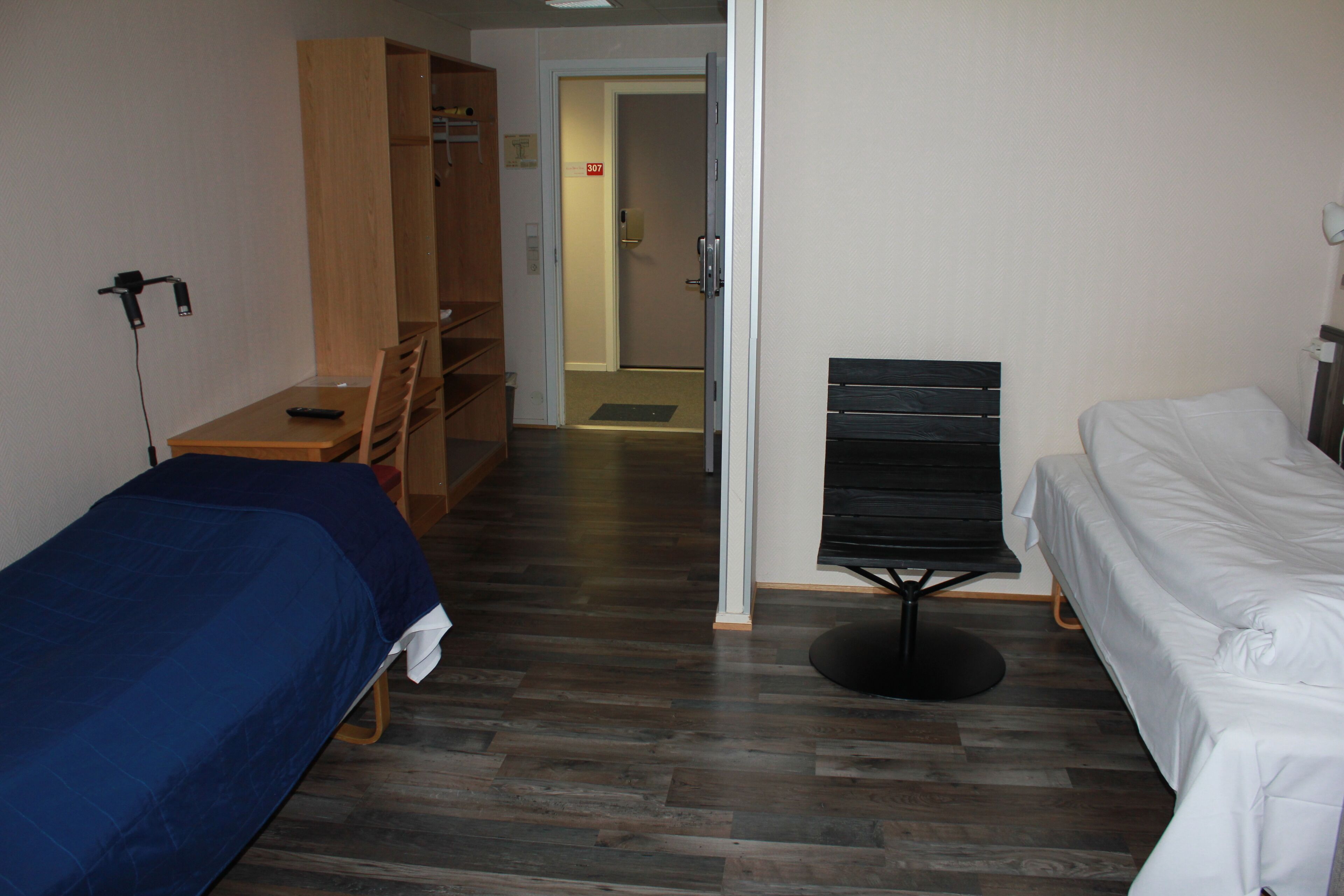 Una habitación de hotel con dos camas, un escritorio, un armario y una puerta que da a un pasillo.