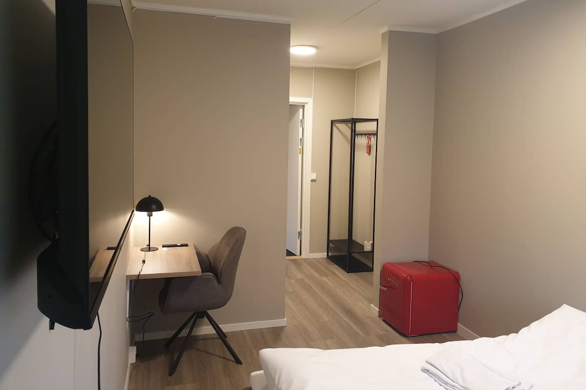 Quarto de hotel com cama, escrivaninha com cadeira, mala vermelha e espelho.