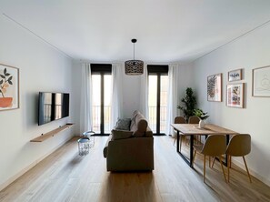 A modern living room with a sofa, a TV, a dining table with chairs, and a large window with curtains. - Apartamento de dos Dormitorios - Picú Home (Málaga)