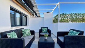 A rooftop patio with a black wicker sofa set, a small table with a plant, and a view of a green fence and clear sky. - Duplex de dos Dormitorios con Terraza - Picú Home (Málaga)