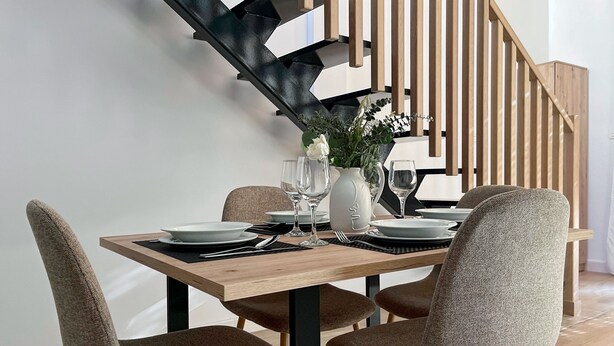 A dining area with a wooden table set for four, surrounded by upholstered chairs. A staircase with wooden railings is visible in the background. - Duplex de dos Dormitorios con Terraza - Picú Home (Málaga)