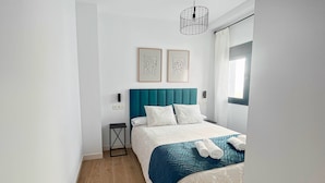 Ein modernes Schlafzimmer mit einem türkisblauen Kopfteil, einem großen Bett mit weißer Bettwäsche, zwei gerahmten Kunstwerken an der Wand und einem kleinen Nachttisch.