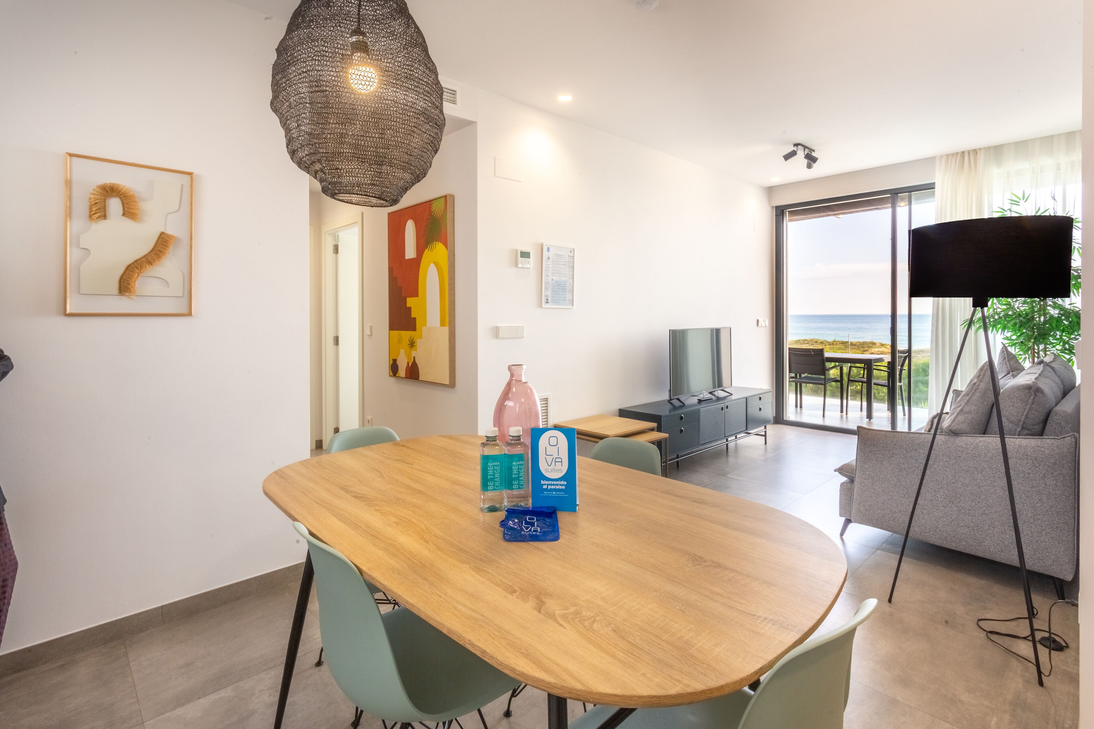 A dining area with a wooden table, chairs, a hanging light fixture, and a view of the ocean through a large window.