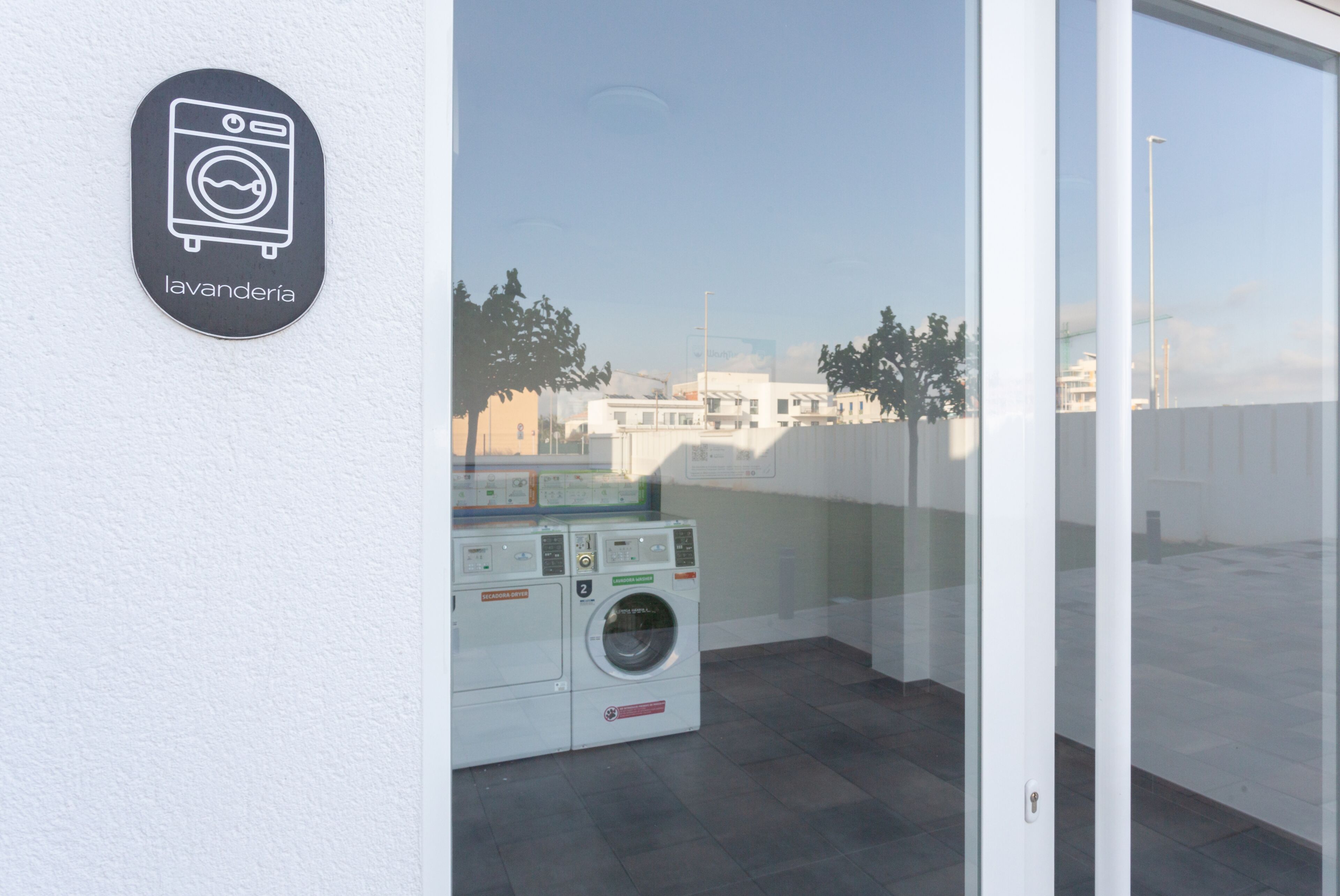 A laundry facility with a washing machine and dryer, a sign indicating 'lavandería,' and a view of buildings through a glass door.