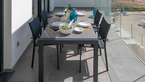 A modern outdoor dining area with a table set for four, large windows, and a view of the sea. - [P2L02] (Oliva)