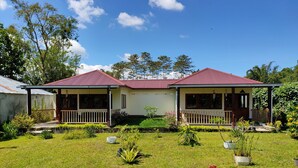 A two-story house with a red roof, a porch with a railing, and a well-maintained garden with various plants and flowers. - ilasan cottage (Tomohon)
