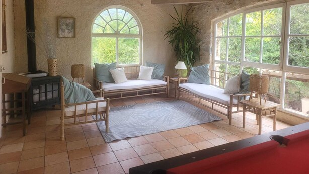 A cozy room with a stone wall, wooden ceiling beams, a large window, and a seating area with a rug and cushions. - Mas "Naturelle" Cévennes (Molières-sur-Cèze)