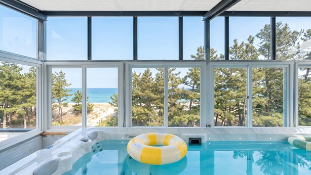 Une piscine avec un anneau gonflable jaune et blanc, de grandes fenêtres donnant sur la mer et les arbres, et un ciel dégagé.