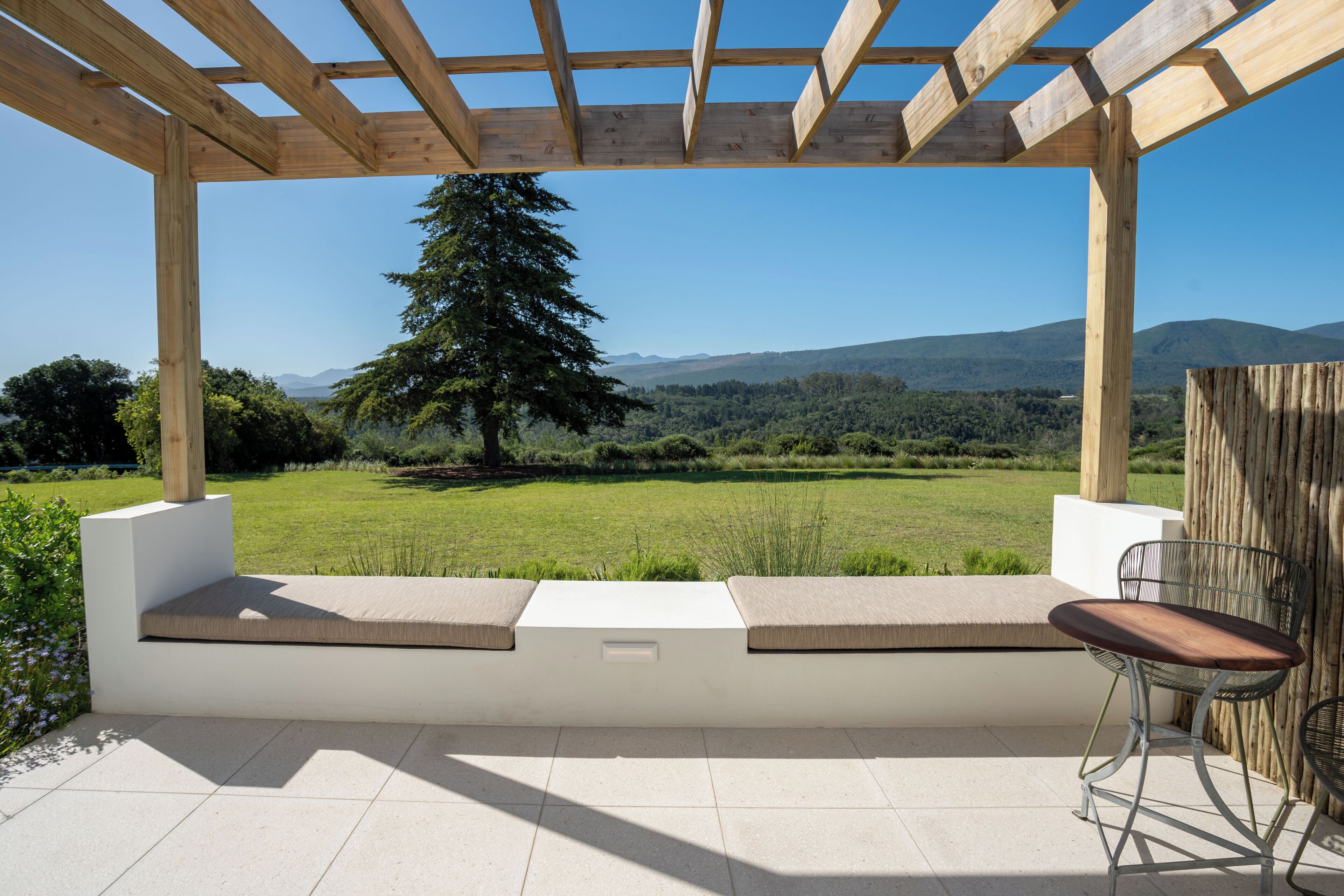A covered outdoor seating area with a bench and chairs, overlooking a grassy landscape and distant hills.