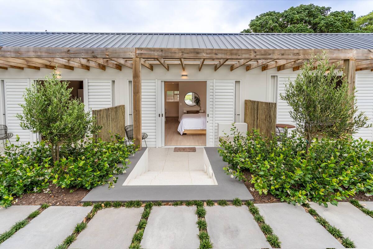 Ein modernes Haus mit einer Holz-Pergola, einem sichtbaren Schlafzimmer mit Bett und Spiegel sowie einem gepflegten Garten mit Sträuchern und einem Weg.