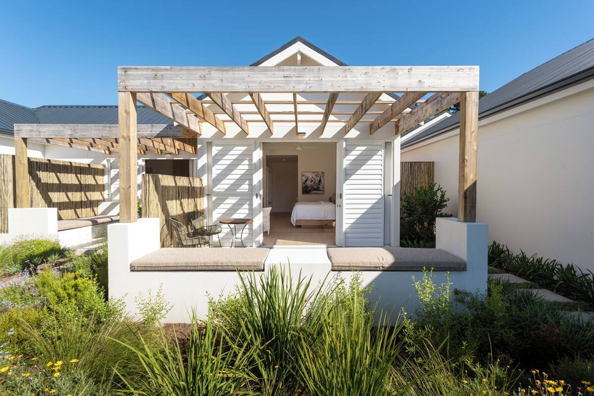 Eine hölzerne Pergola mit Liege und Sitzgelegenheit darunter, umgeben von einem Garten mit verschiedenen Pflanzen.