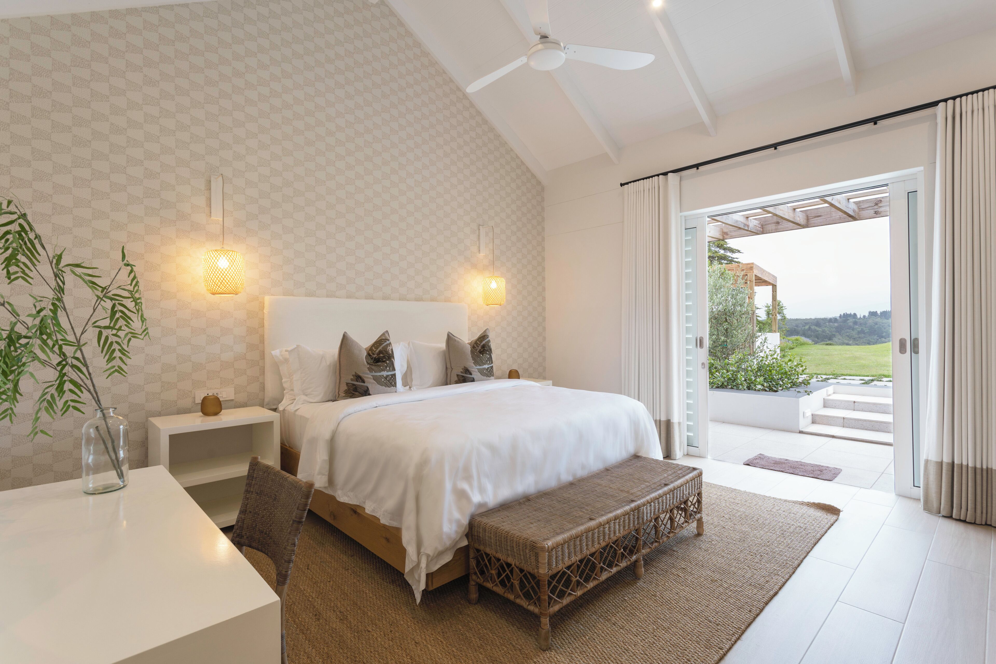 A bedroom with a large bed, bedside tables, a bench, and a view of the outdoors.