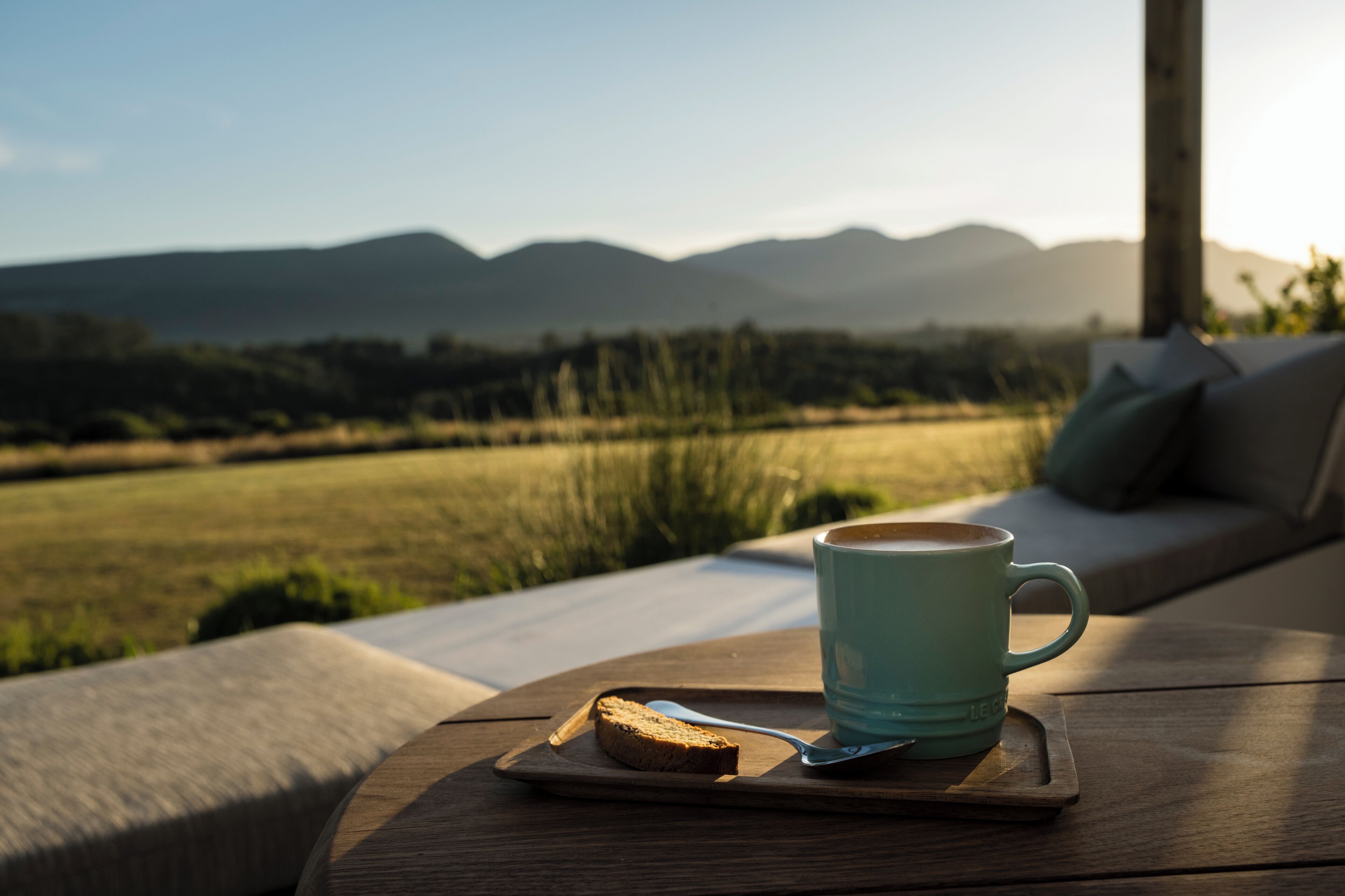 Ein Holztisch mit einer Kaffeetasse und einer Brotscheibe auf einem Tablett, mit Blick auf ein Feld und Berge.