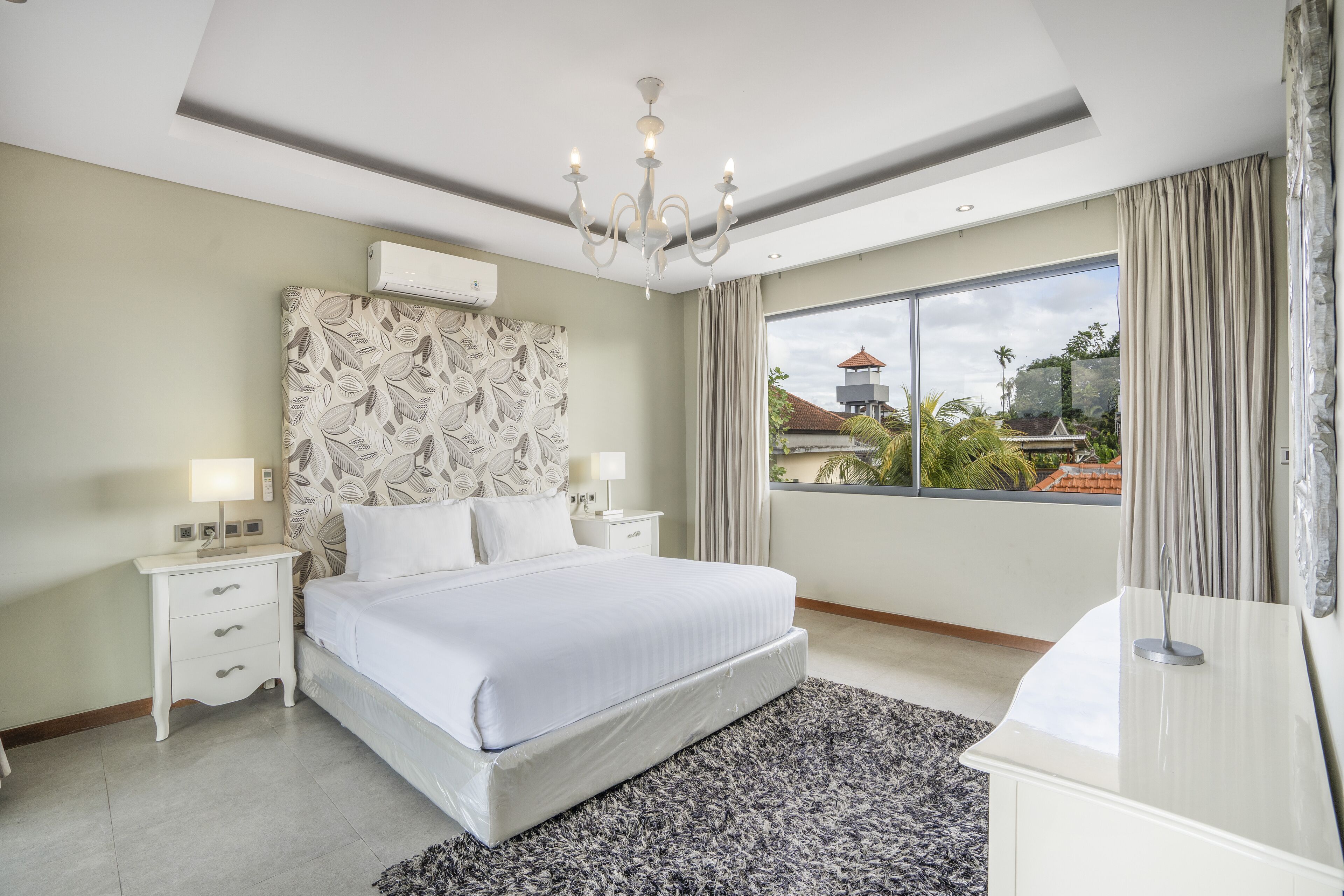 A modern bedroom with a large bed, a chandelier, a view of the outside, and a nightstand with a lamp.