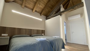 Une chambre avec deux lits, des poutres apparentes en bois au plafond, une porte blanche et une table de chevet avec une lampe.