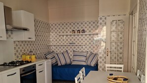A compact kitchen with white cabinets, a gas stove, and a built-in oven. There is a blue sofa with striped cushions and a dining table with white chairs. - Luna apartment with free parking. (nervi genova)